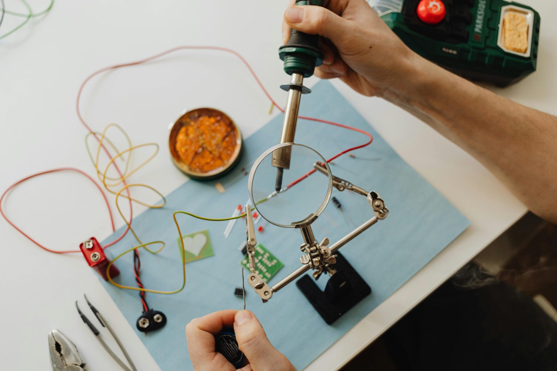 Hände löten aus großer Höhe mithilfe einer Lupe und Werkzeugen eine Leiterplatte.